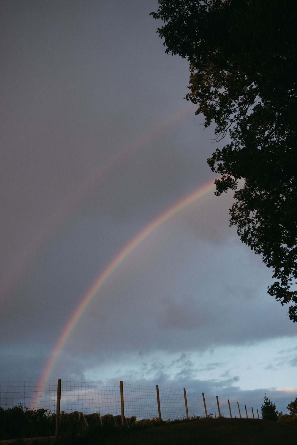 Double rainbow arches over a rural fence line at dusk with dark storm clouds in the background.