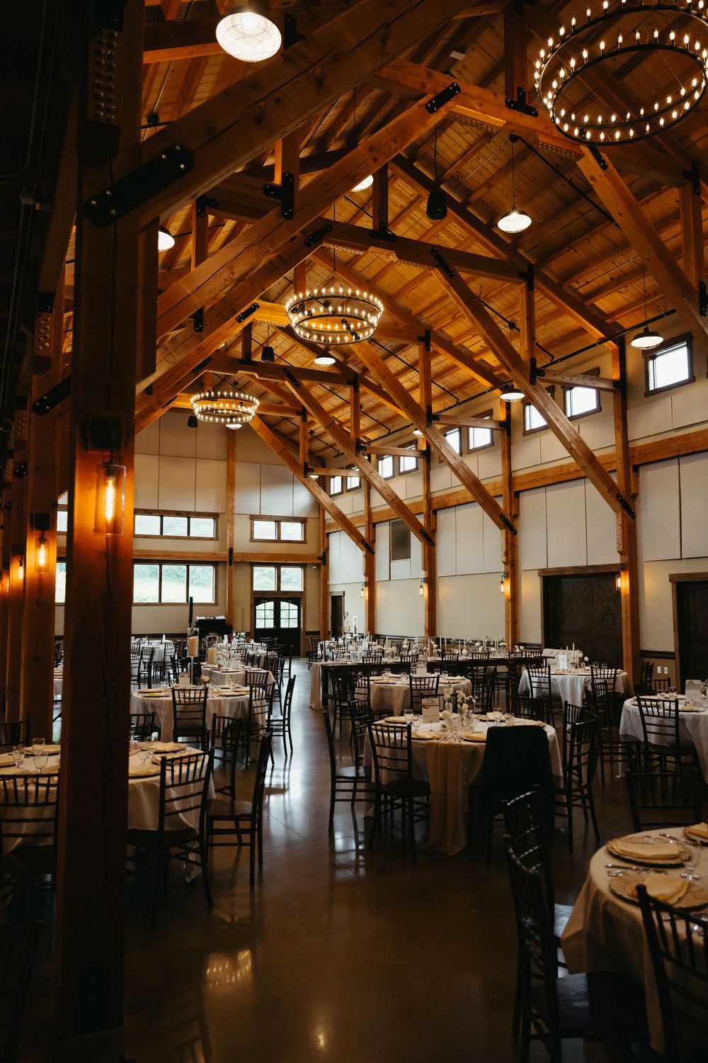 Rustic wooden barn venue interior with elegant chandeliers and dark chiavari chairs set up for an elegant reception.