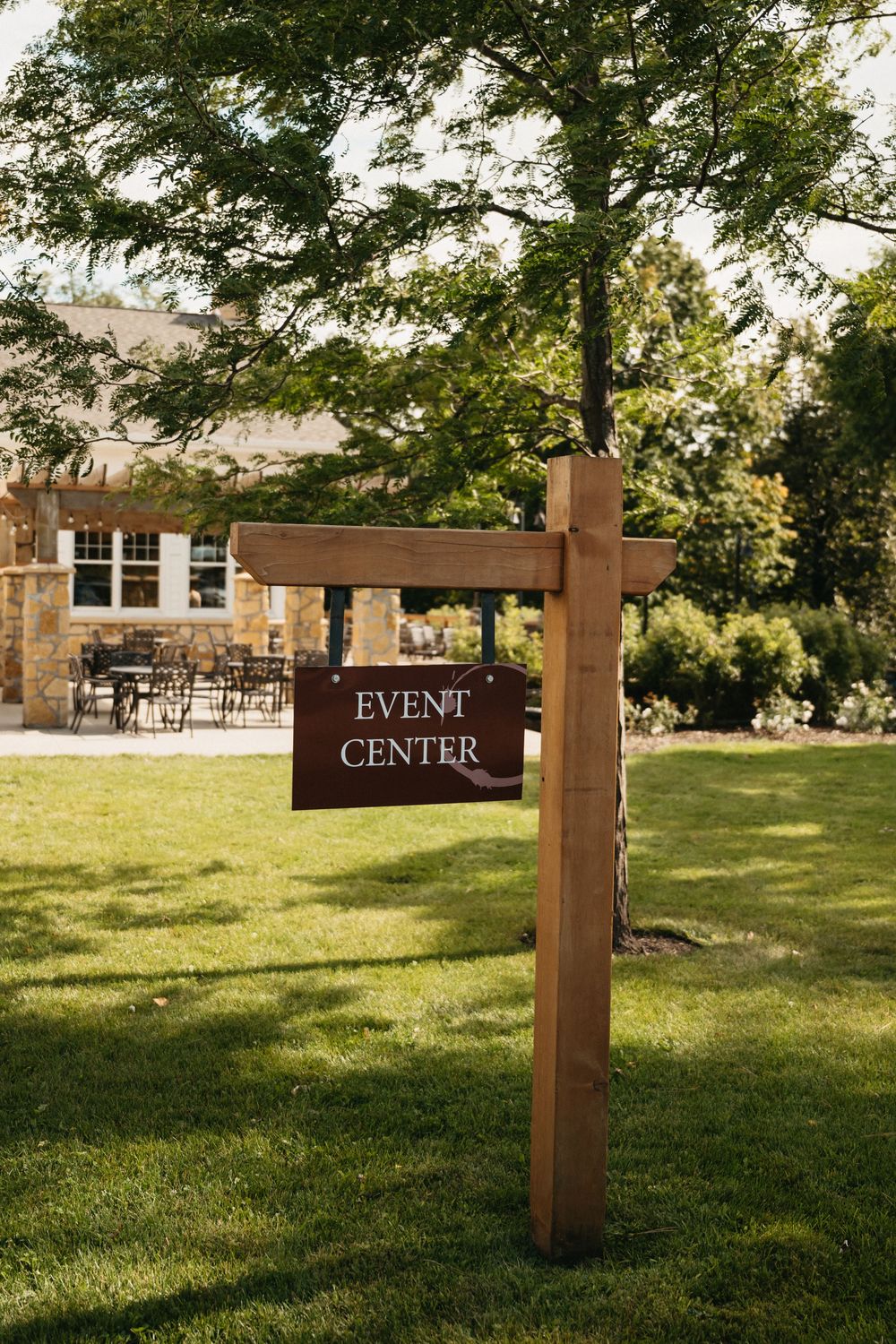 Wooden directional sign for 'Event Center' stands in a sunny green lawn with trees and buildings in the background.