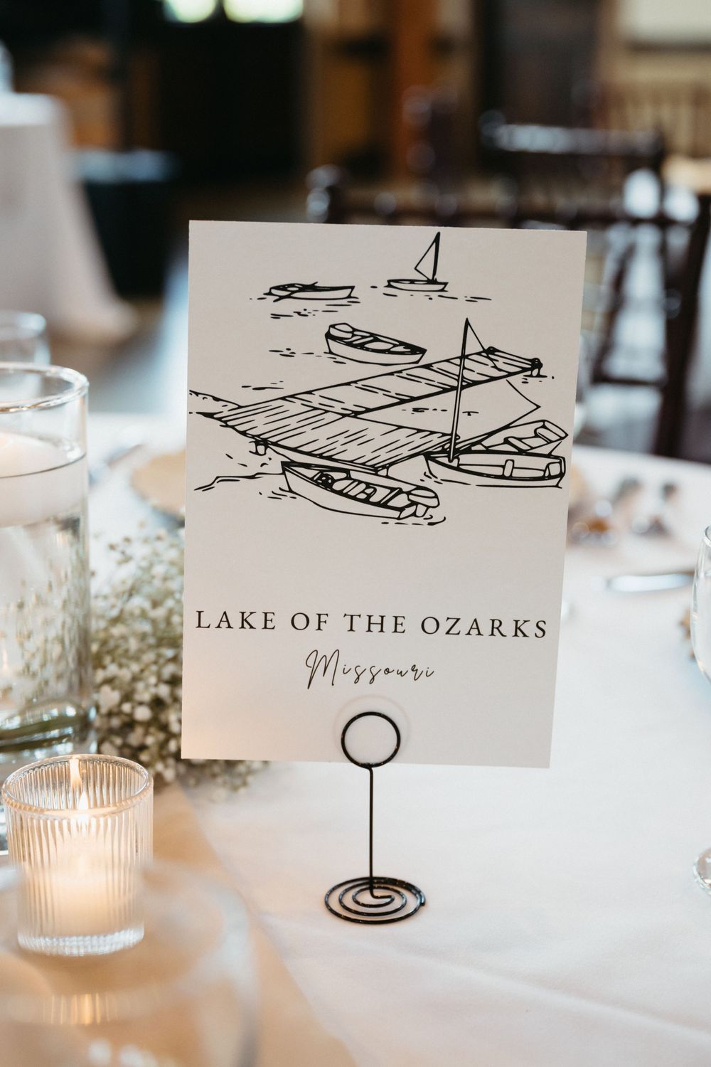 Wedding reception table cards feature elegant sketches of boats and Lake of the Ozarks destination names with candlelit decor.