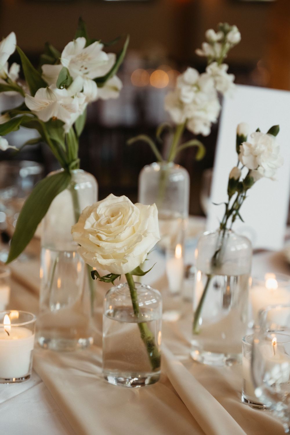 White roses and lilies in glass vases line an elegant table setting with glowing candles.