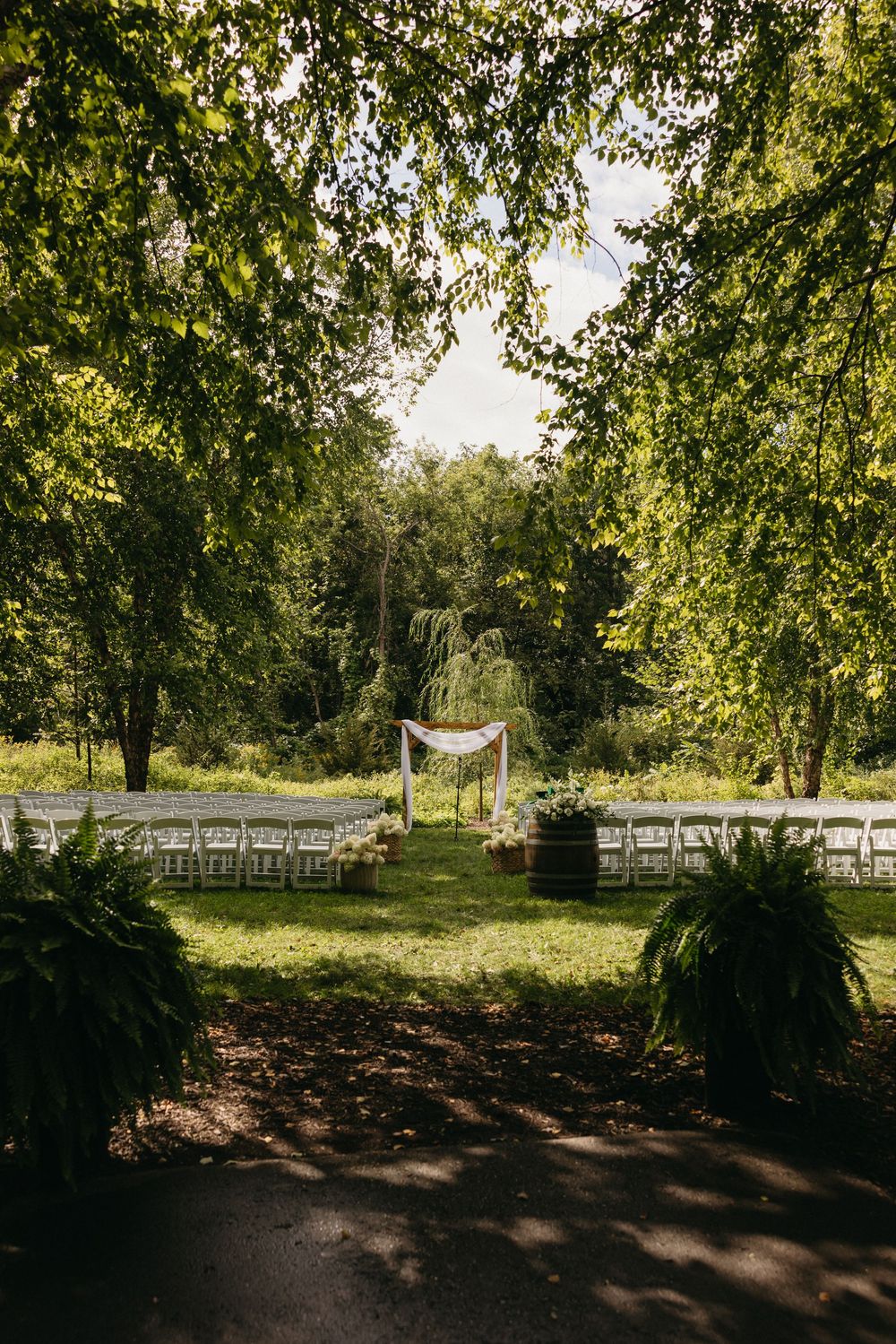 Outdoor wedding ceremony setup with white chairs arranged in rows along a tree-lined path in natural sunlight.