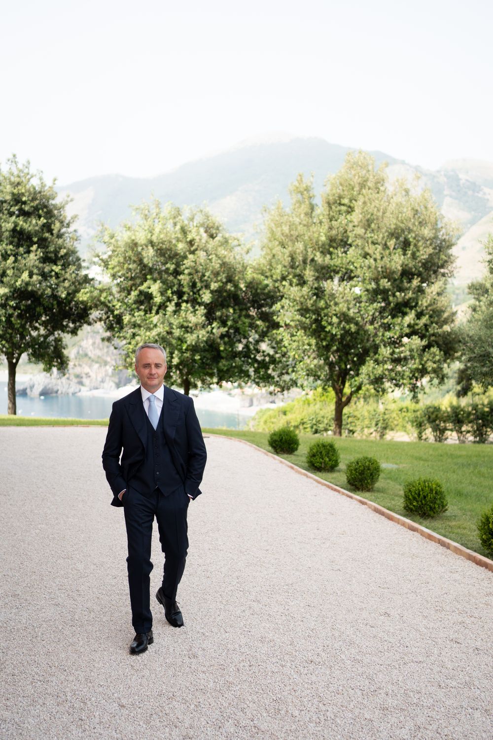 A person in a dark suit walks along a gravel path lined with trees and mountains in the background.