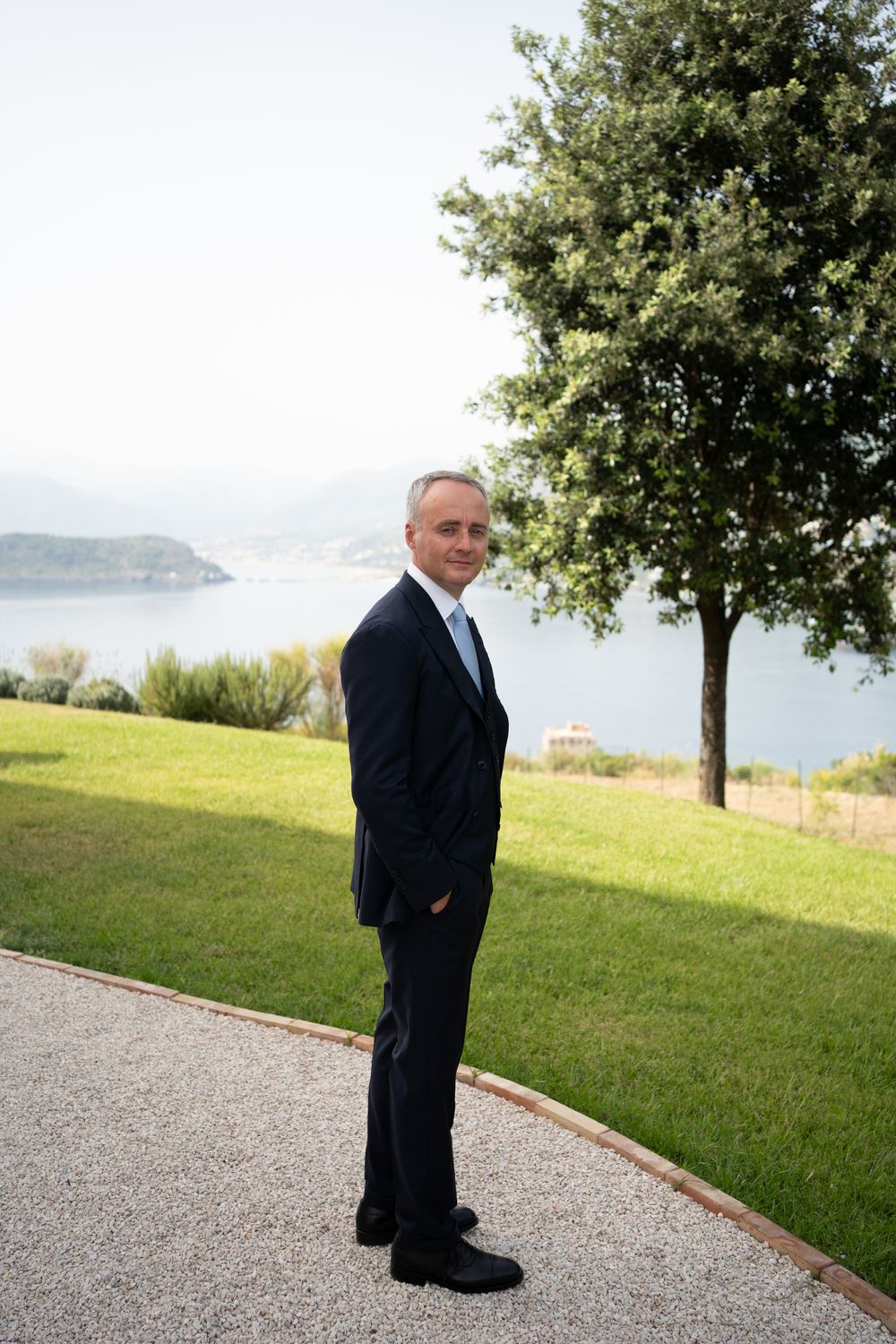 A figure in a dark suit stands on a grassy hillside overlooking a scenic coastal landscape.