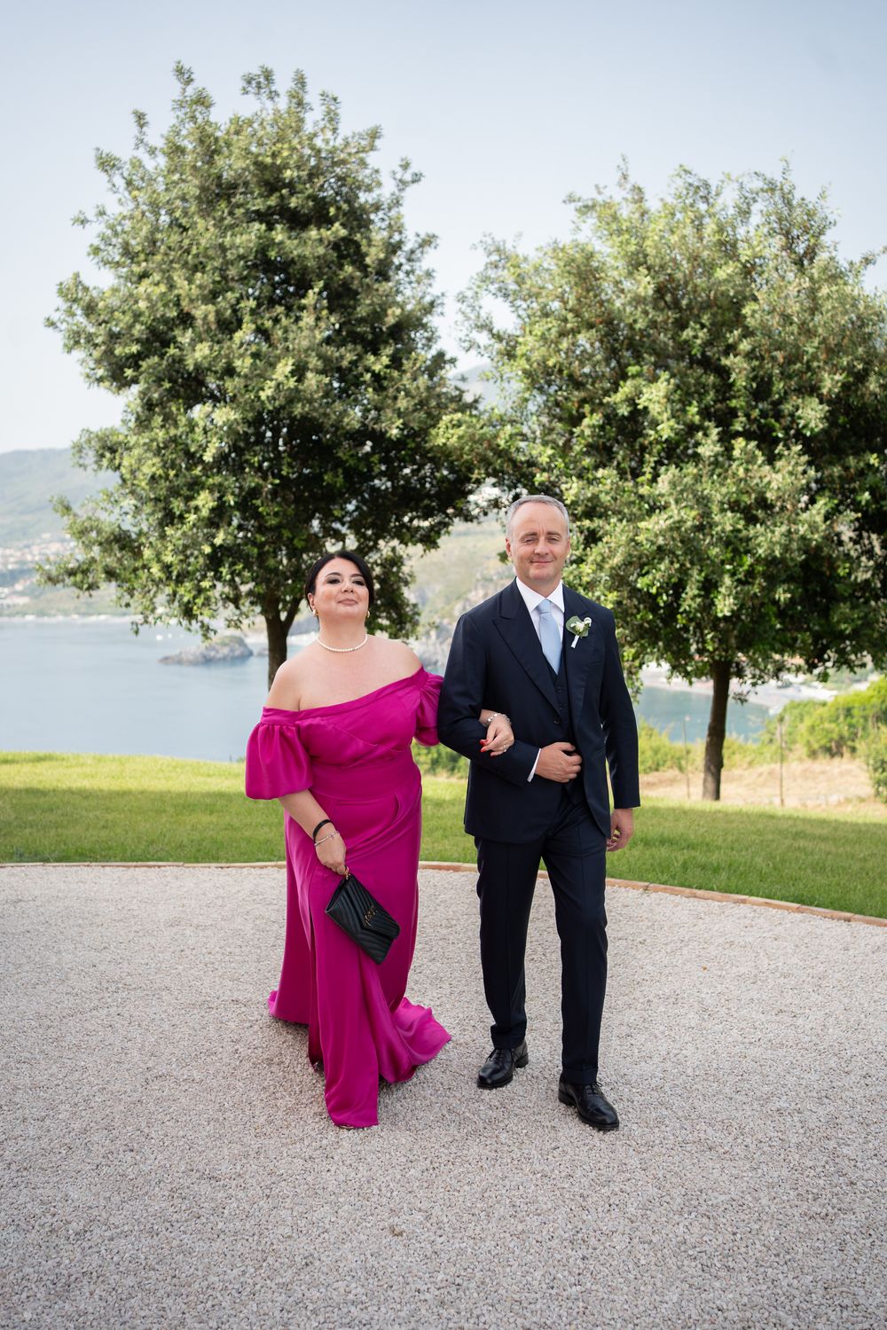 A couple walks together outdoors, with the woman wearing a vibrant pink dress and the man in a dark suit.