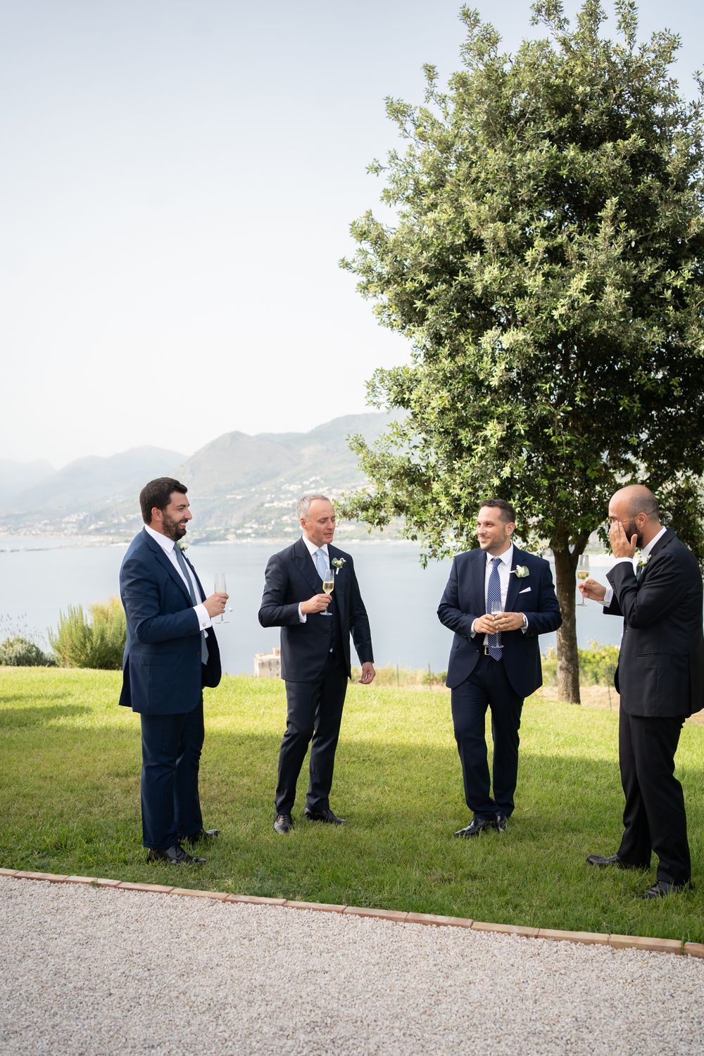 Groomsmen in navy suits chatting and laughing on a lawn with mountain and water views.