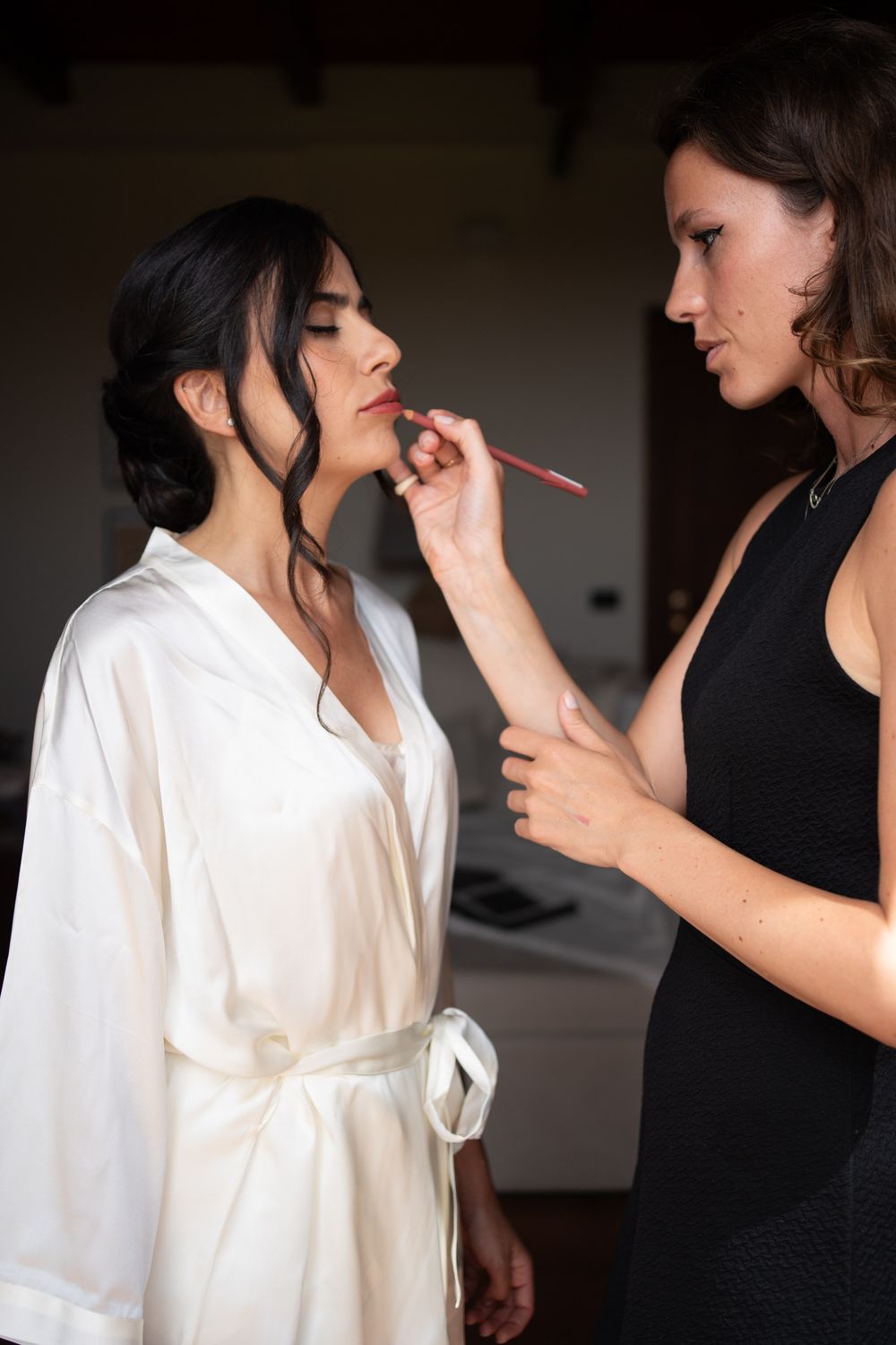 Makeup artist applying cosmetics to bride in white robe during wedding preparation.