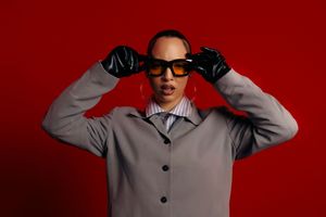 Person in a gray sweater poses against a red background while adjusting their sunglasses.