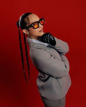 A person in a gray hoodie and sunglasses poses against a deep red background in a stylish fashion photoshoot.