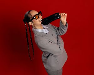 Person in sunglasses and grey coat posing dramatically against a red background.