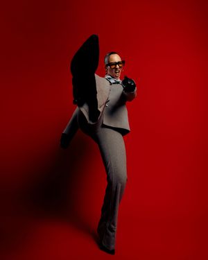 Figure in a gray suit and glasses strikes a dramatic pose against a deep red background.