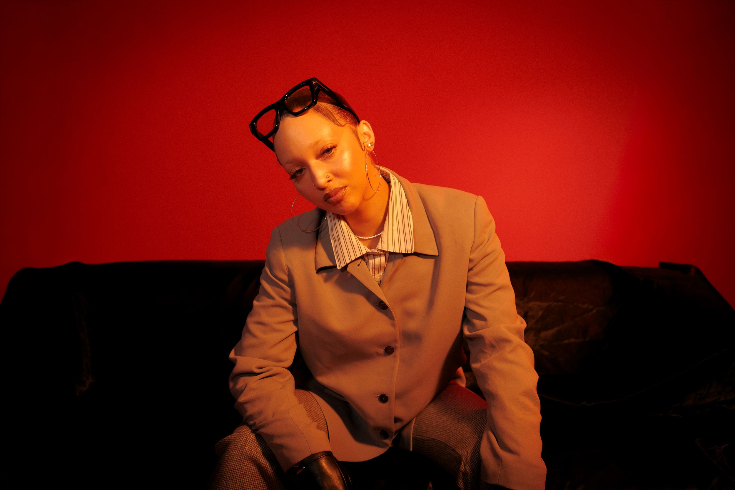 Figure in a beige coat sits on dark couch against dramatic red backdrop with moody lighting.