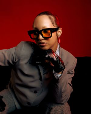Person in sunglasses and grey suit jacket poses against dark red background in artistic photo series.