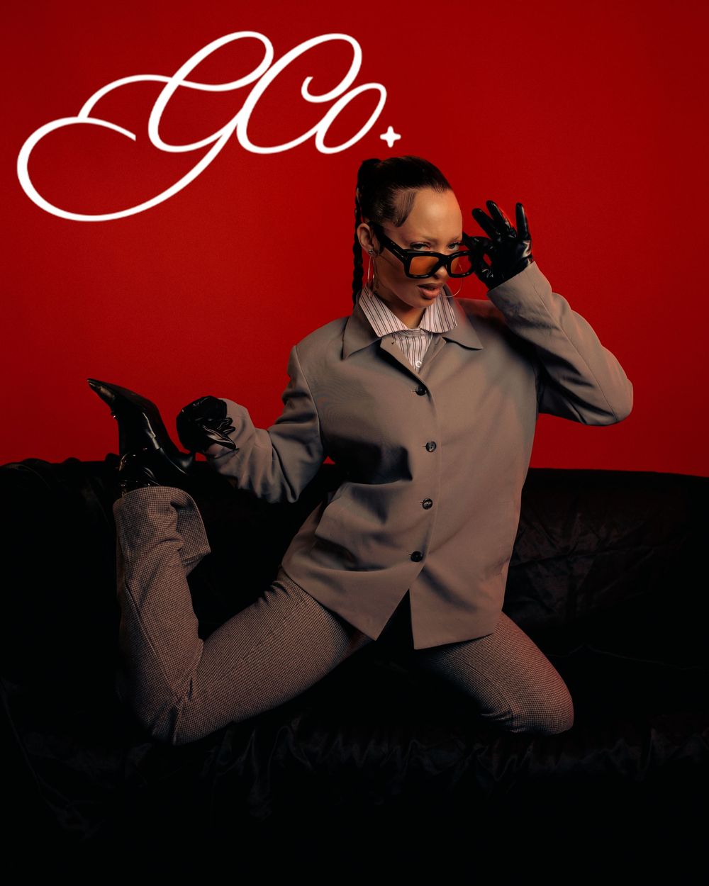 A model poses on a dark sofa in a gray suit with black gloves and heels against a bold red background with GCo logo.