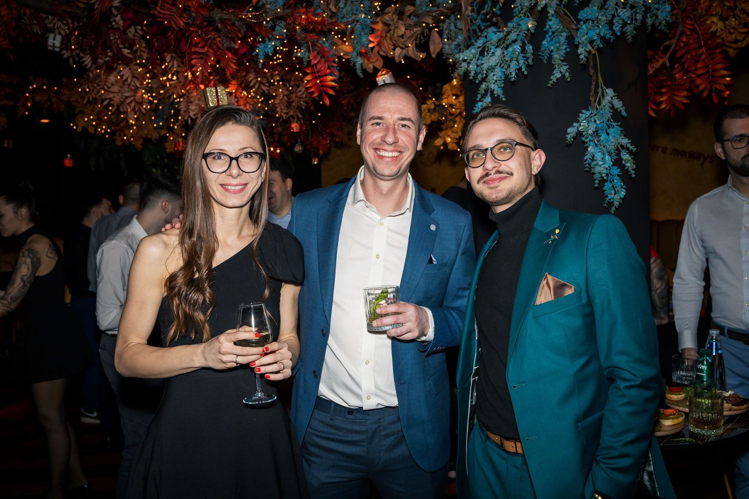 Two men and a woman posing with cocktails at festive indoor party with floral decorations.