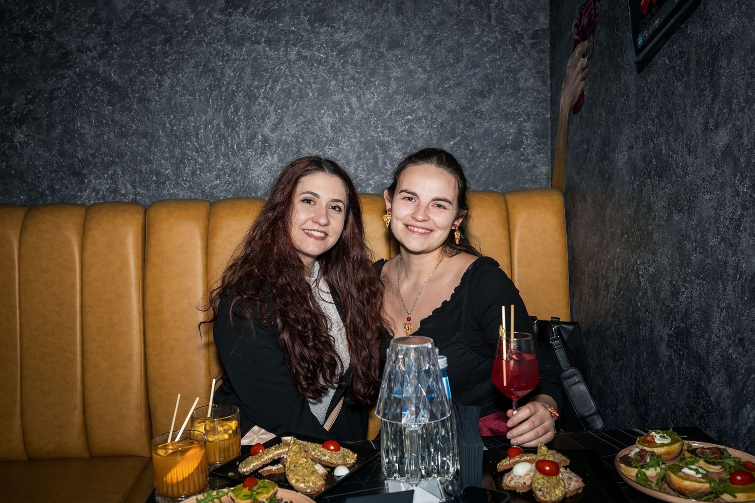 Two young women smiling together at restaurant booth with food and cocktails on table.