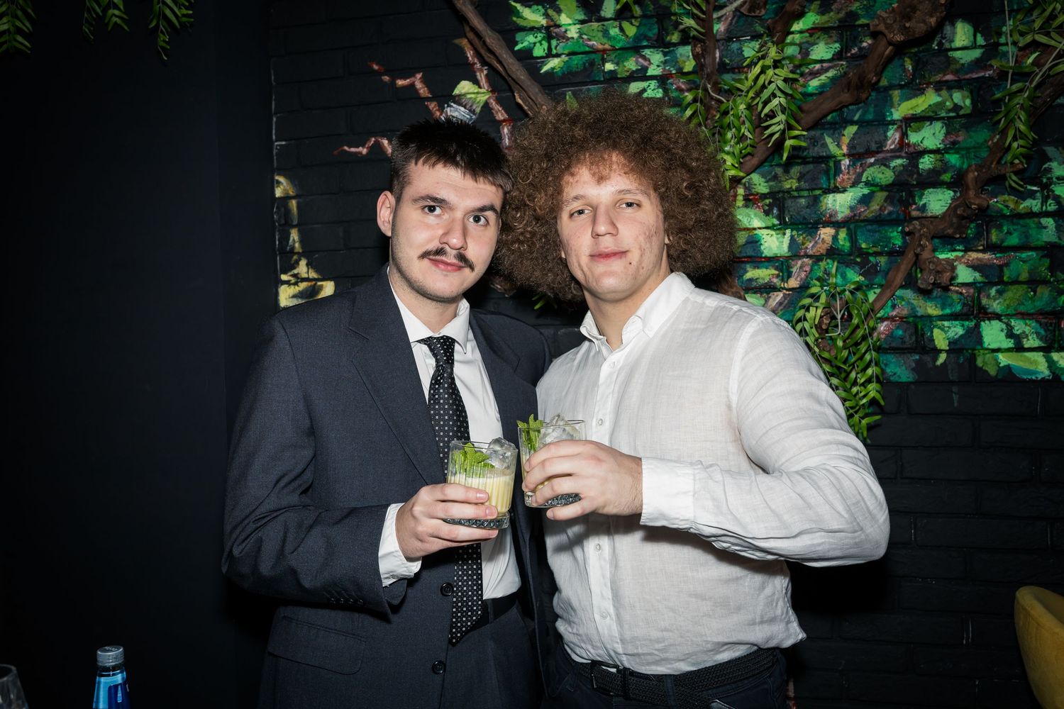 Two young men holding mojito cocktails posing together at indoor party event.