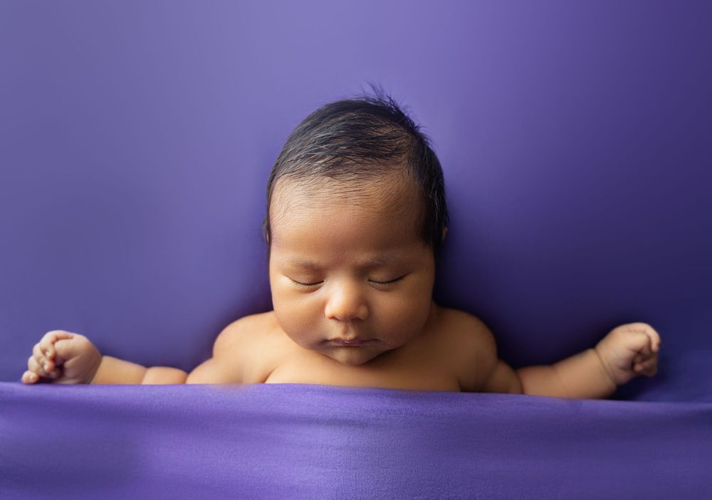 Newborn baby resting chin on hands against a purple fabric background.