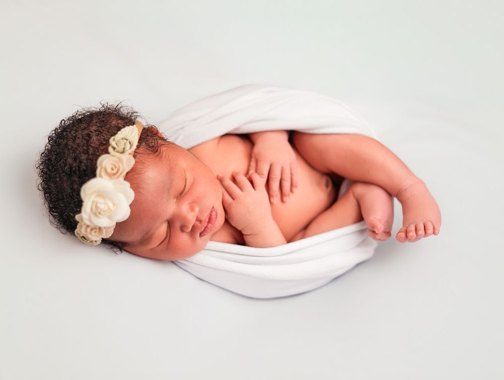Newborn baby girl sleeping peacefully wrapped in white cloth with floral headband.