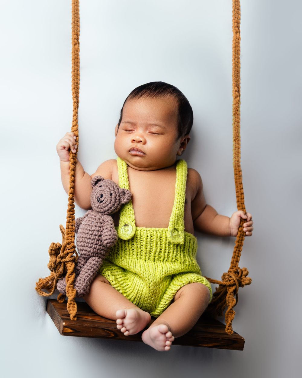 Sleeping newborn baby in green knit romper holding teddy bear on wooden swing with rope.