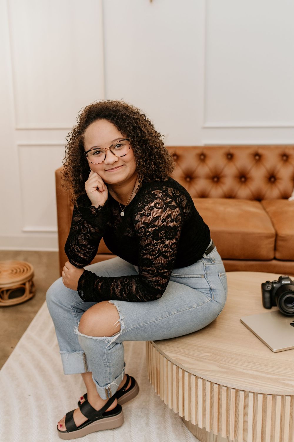 Portrait of person in lace top and ripped jeans sitting on brown leather furniture.