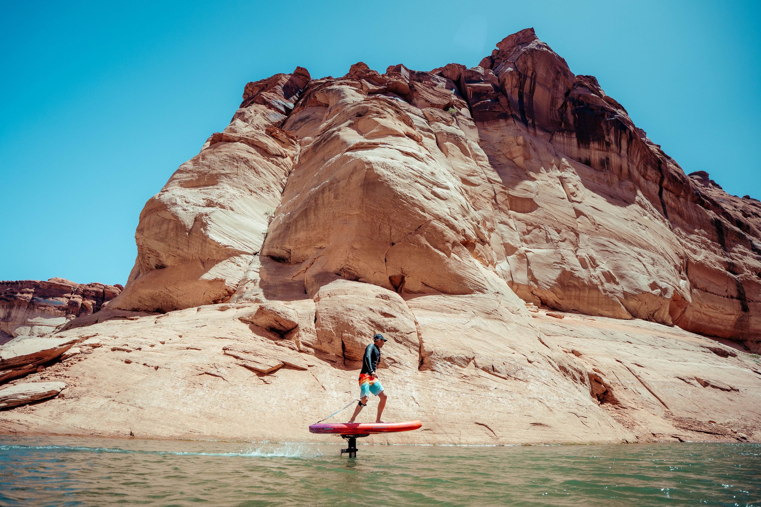 Stand up paddleboarder glides past towering red rock formations at Lake Powell on a sunny day.