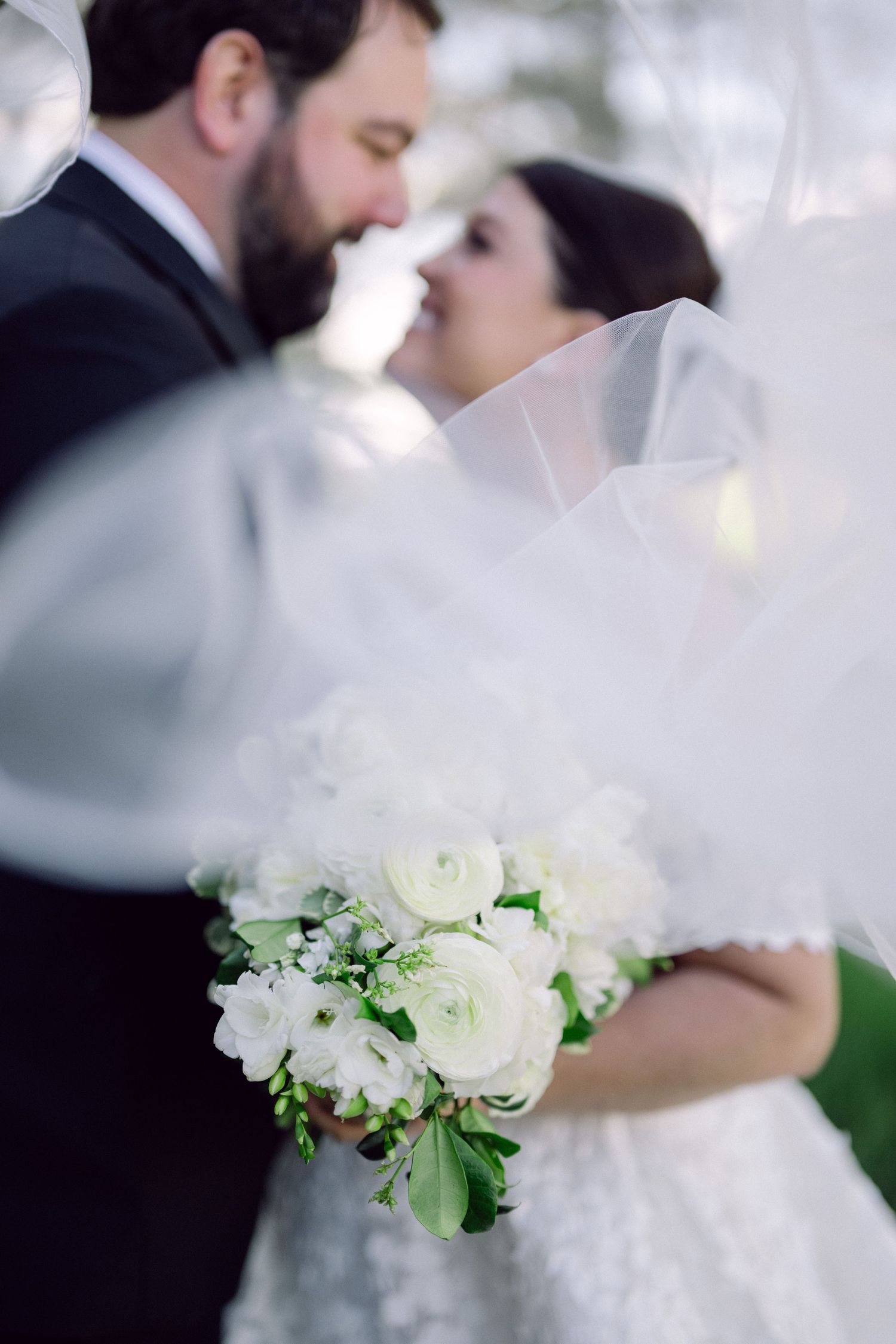 A romantic wedding moment captured with a soft-focus white bouquet in the foreground and a flowing veil.
