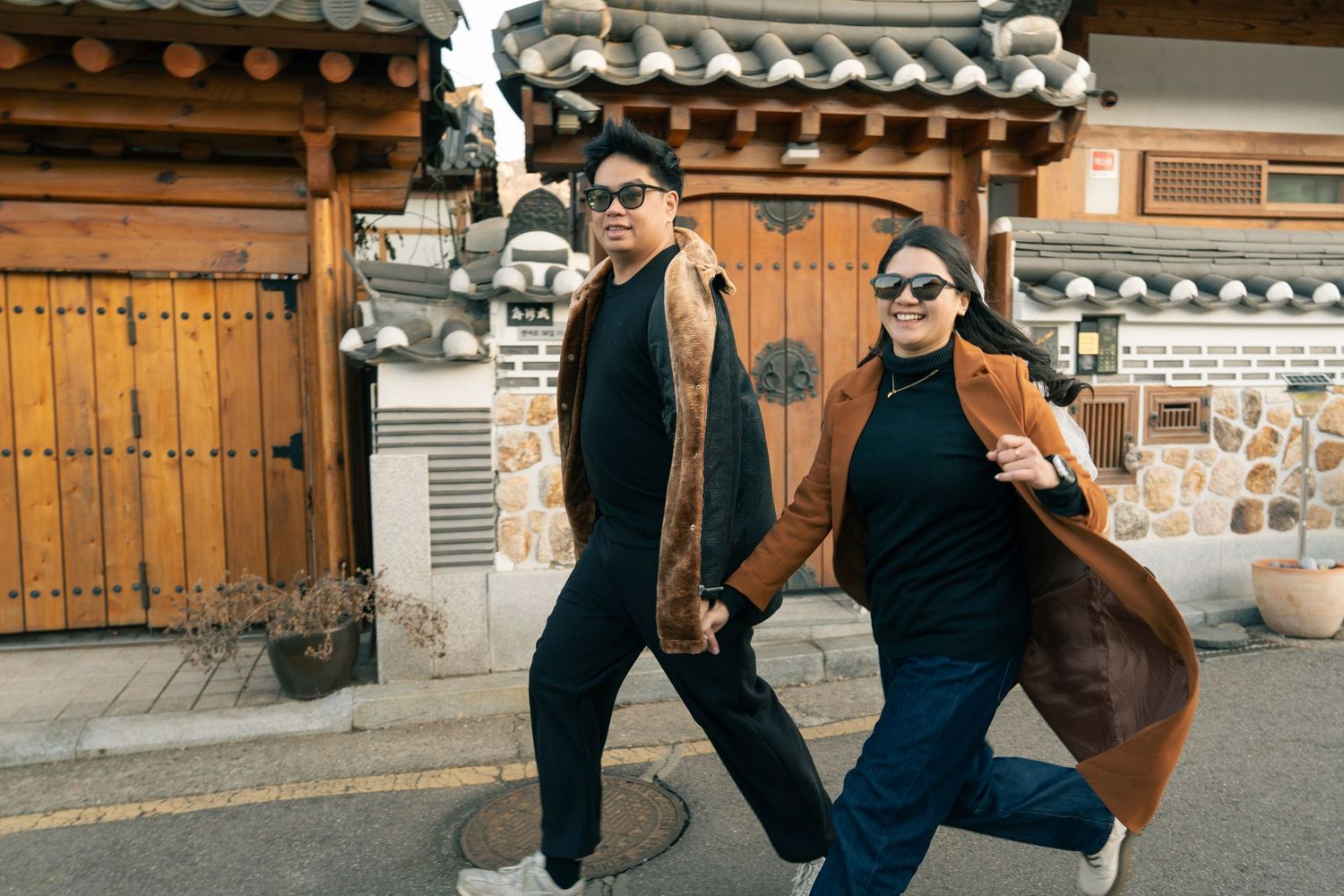 Two people in casual attire pose playfully in front of traditional Korean architecture and wooden doors.