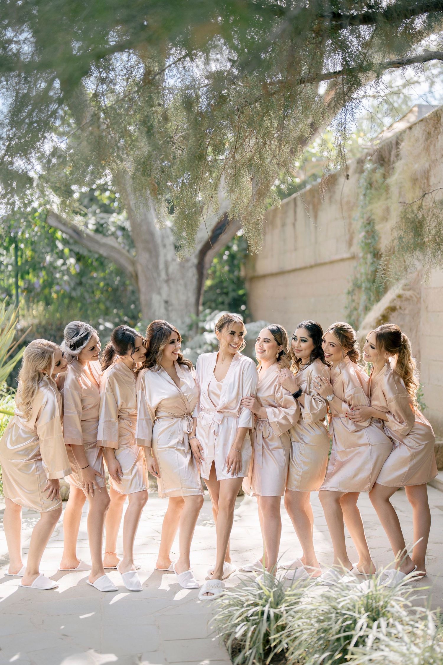 A group in matching blush pink robes pose in a line against a garden wall, creating a playful bridal party photo.