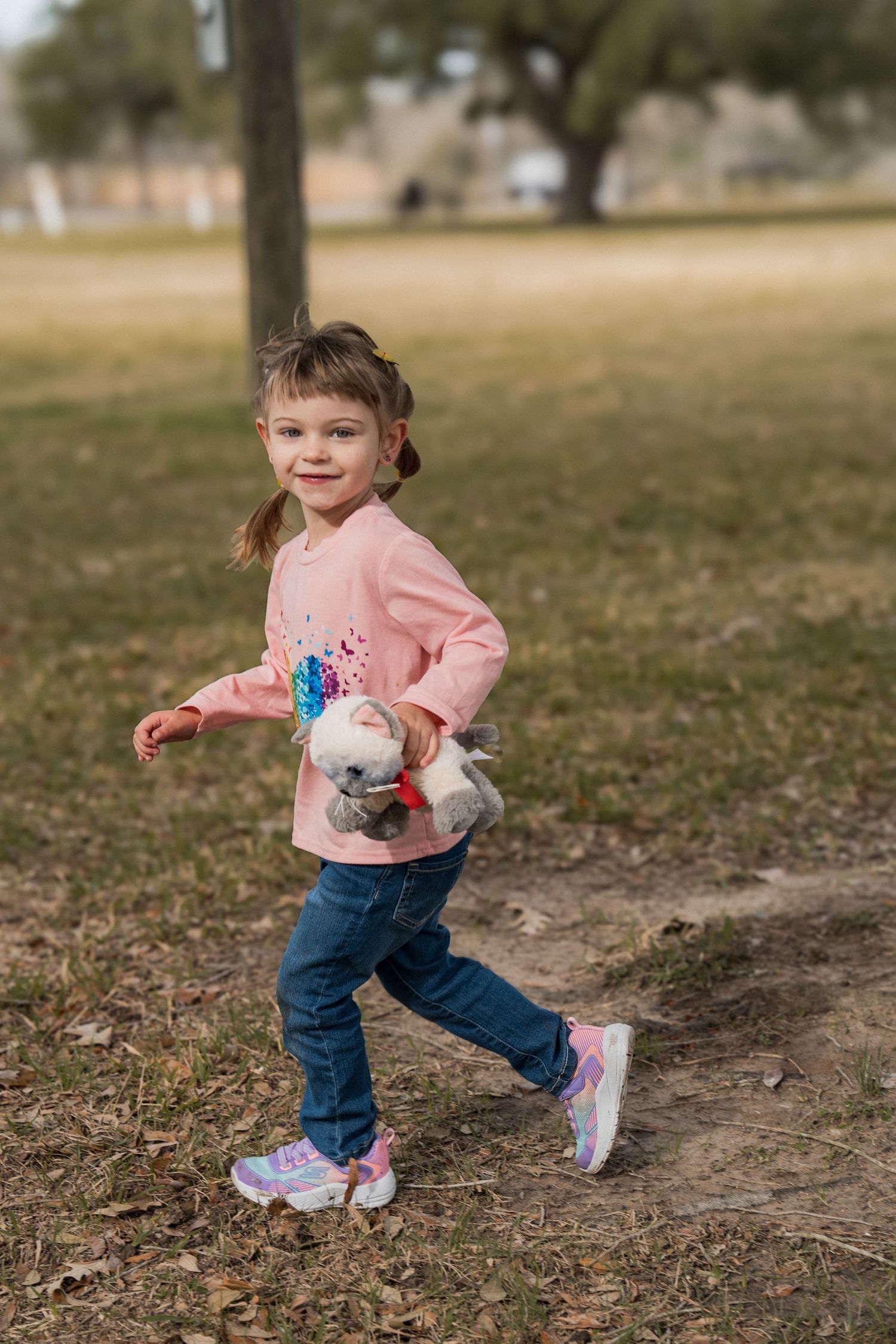 A small child wearing a pink sweater and jeans runs playfully through a park on a sunny day.