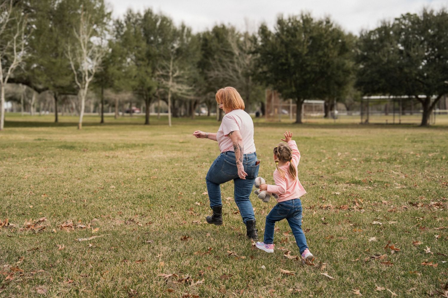 Two people run and play together in a grassy park area with trees in the background.
