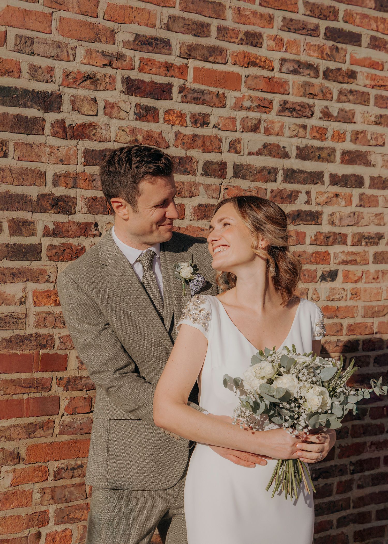 Romantic wedding portrait against brick wall with bride holding white bouquet.