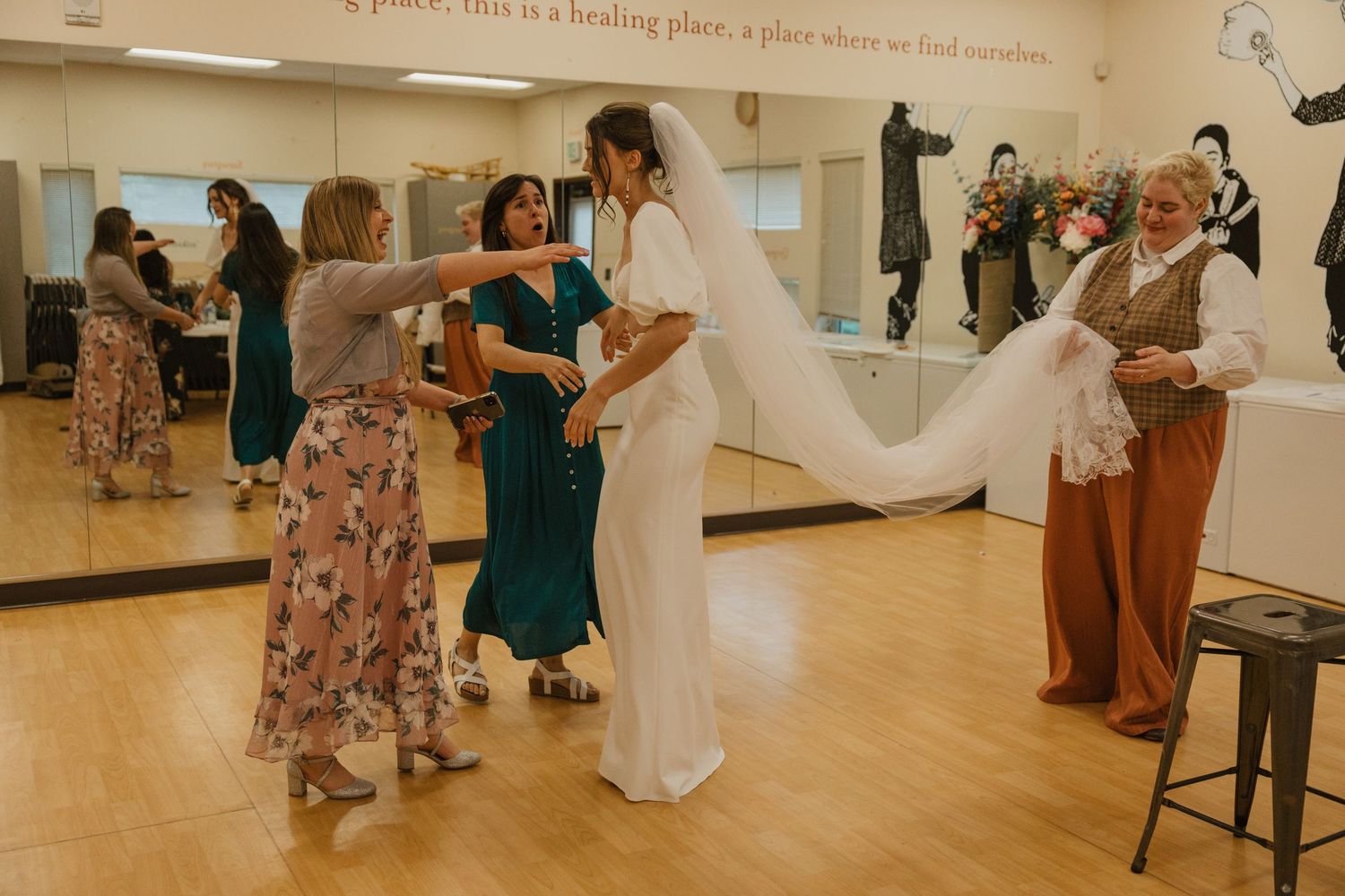 Bride in white gown being helped by bridesmaids with her veil in a bright wedding prep room.
