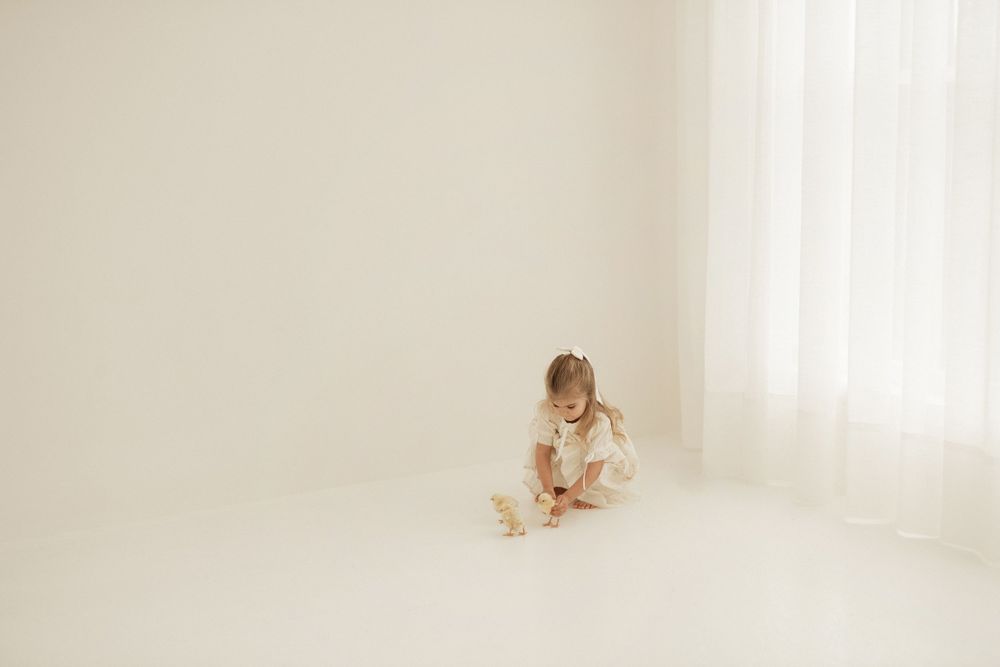 A small child in a white dress sits reading in a bright minimalist corner of a room with soft natural light.