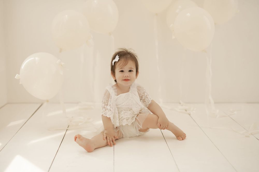 A baby in a white lace dress sits and plays on a light-colored floor in a dreamy, ethereal setting.