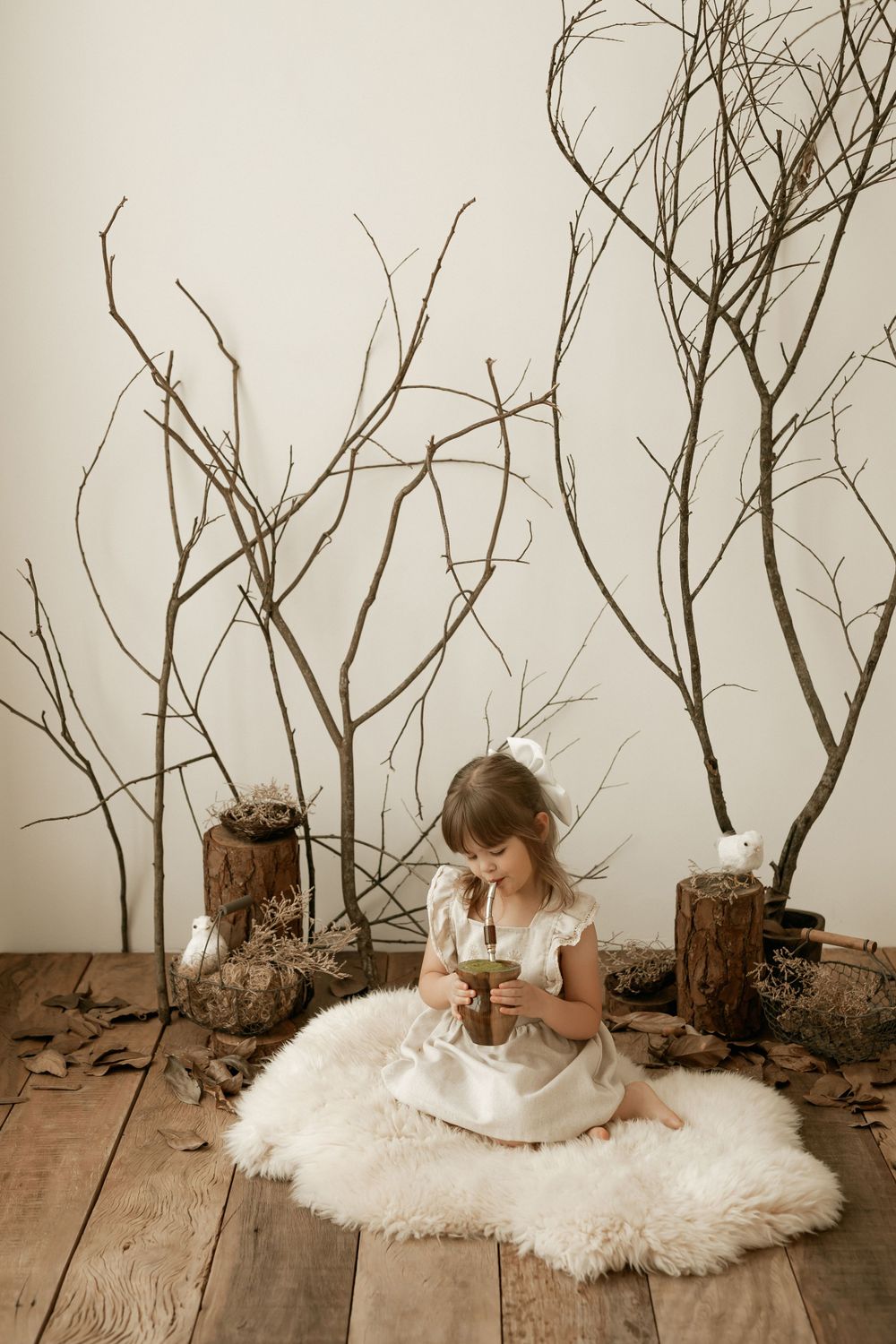Young child in white dress sits on fluffy rug amid bare branches and wooden stumps in rustic indoor setting.