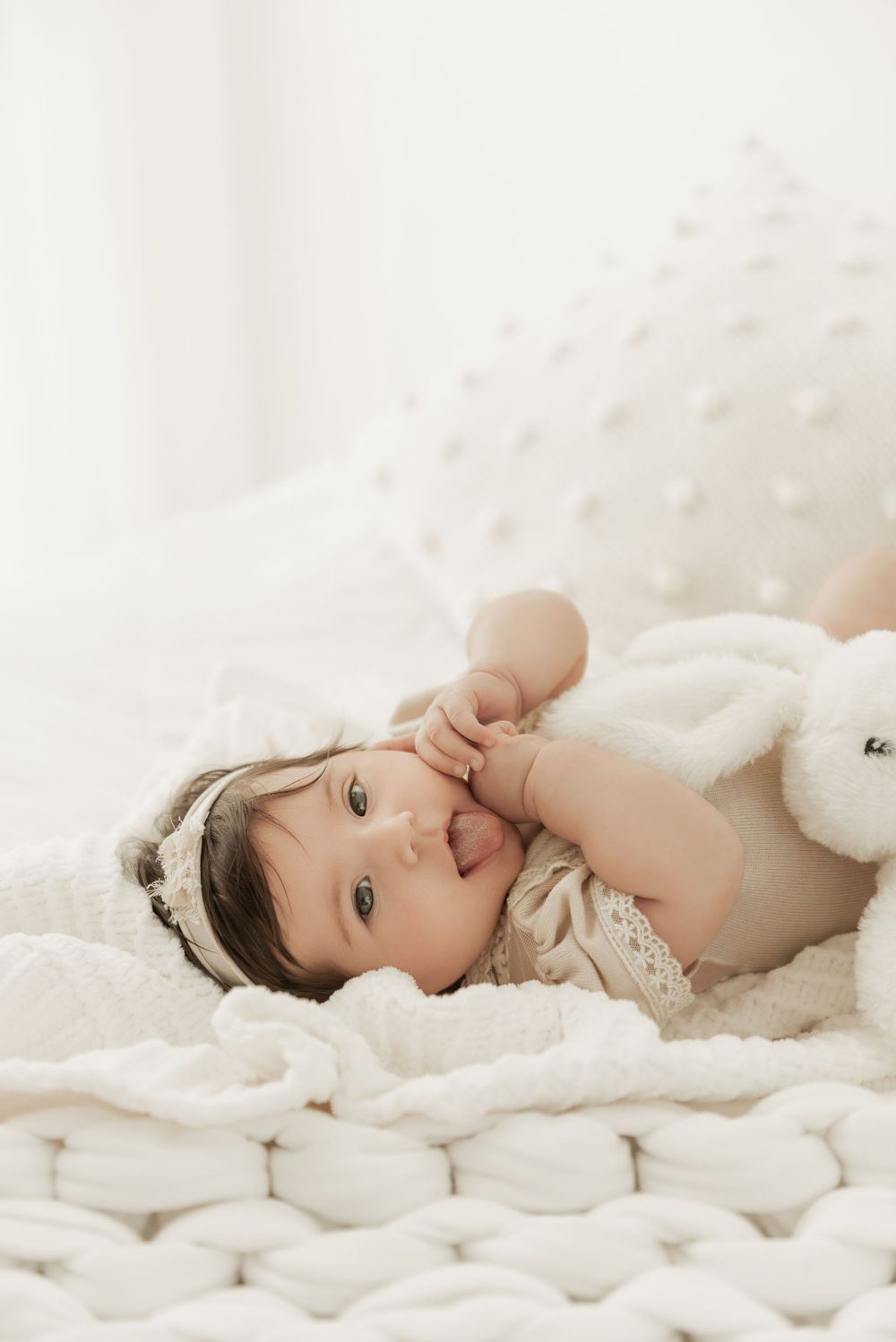 A baby lies peacefully on soft white textured bedding in a dreamy natural light setting.