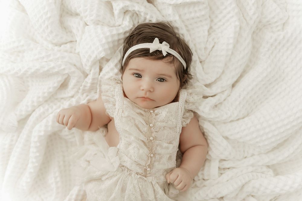 A baby wearing a white lace dress and headband lies on soft white textured fabric in a dreamy portrait setting.
