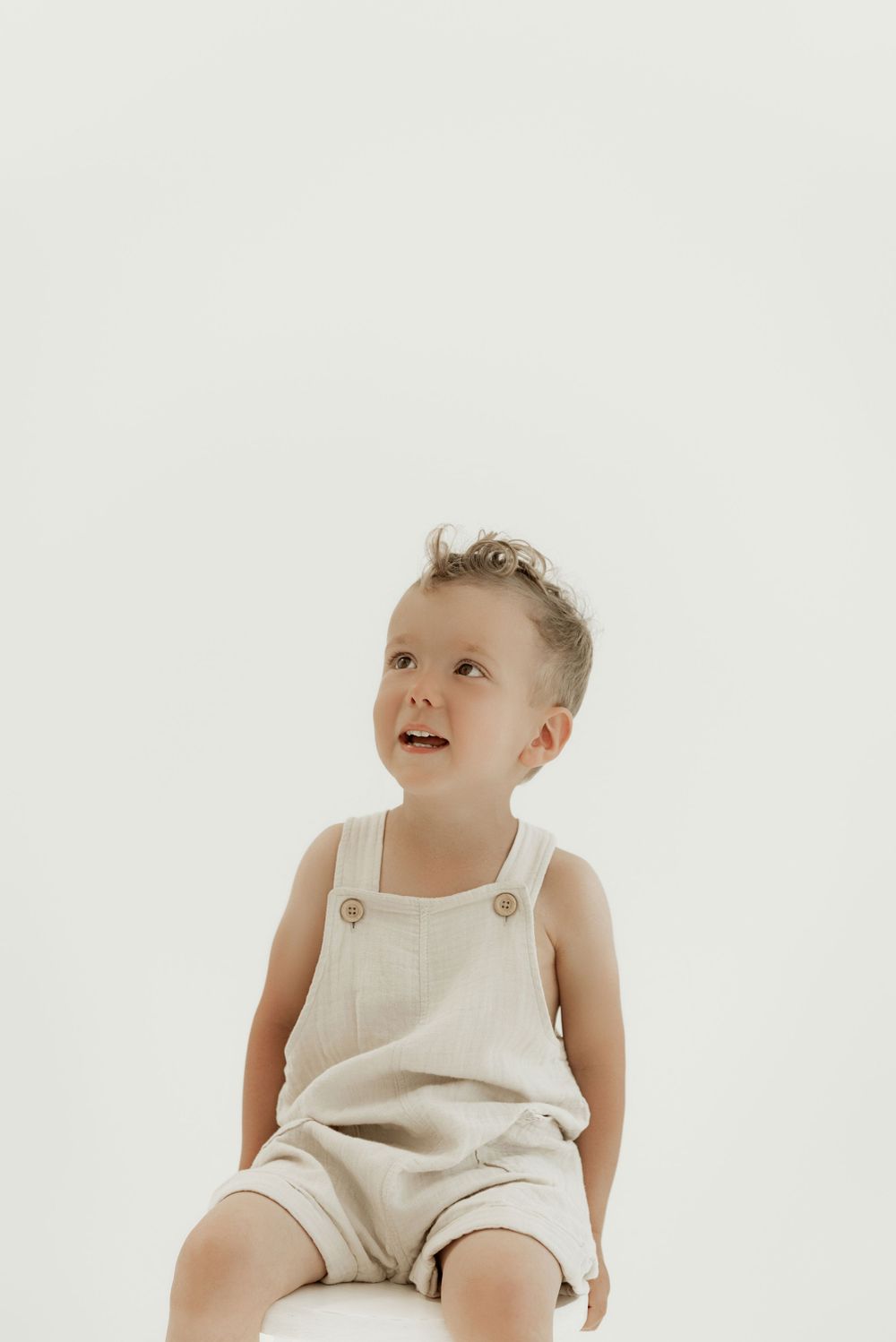 A small child wearing a cream-colored linen romper sits and smiles against a white background.