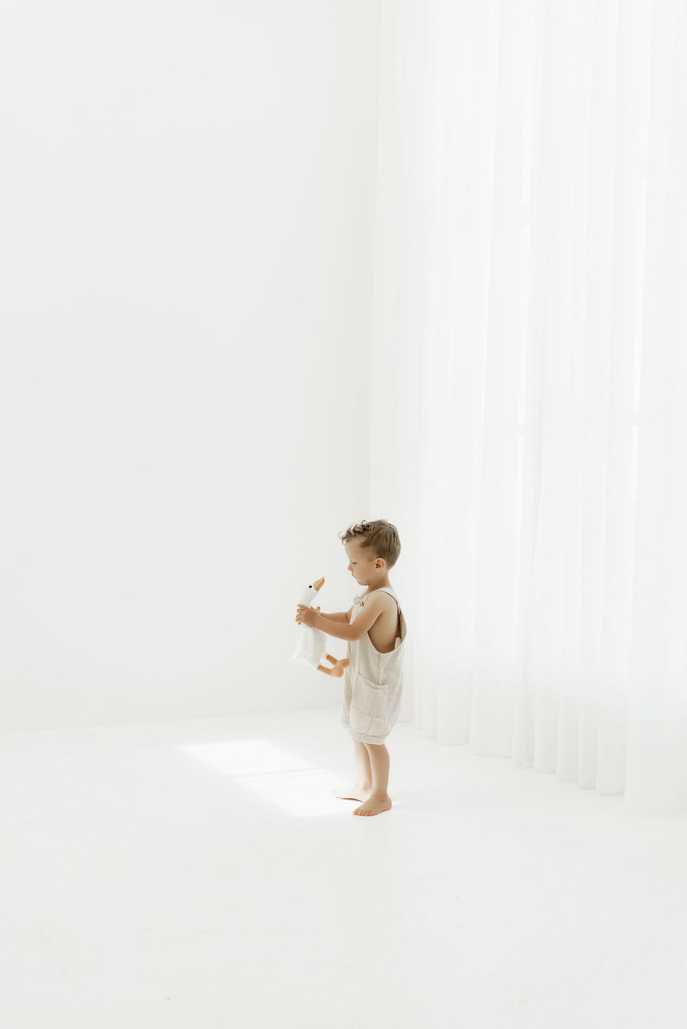 A small toddler in a white dress stands against a bright white background, creating a minimalist artistic portrait.