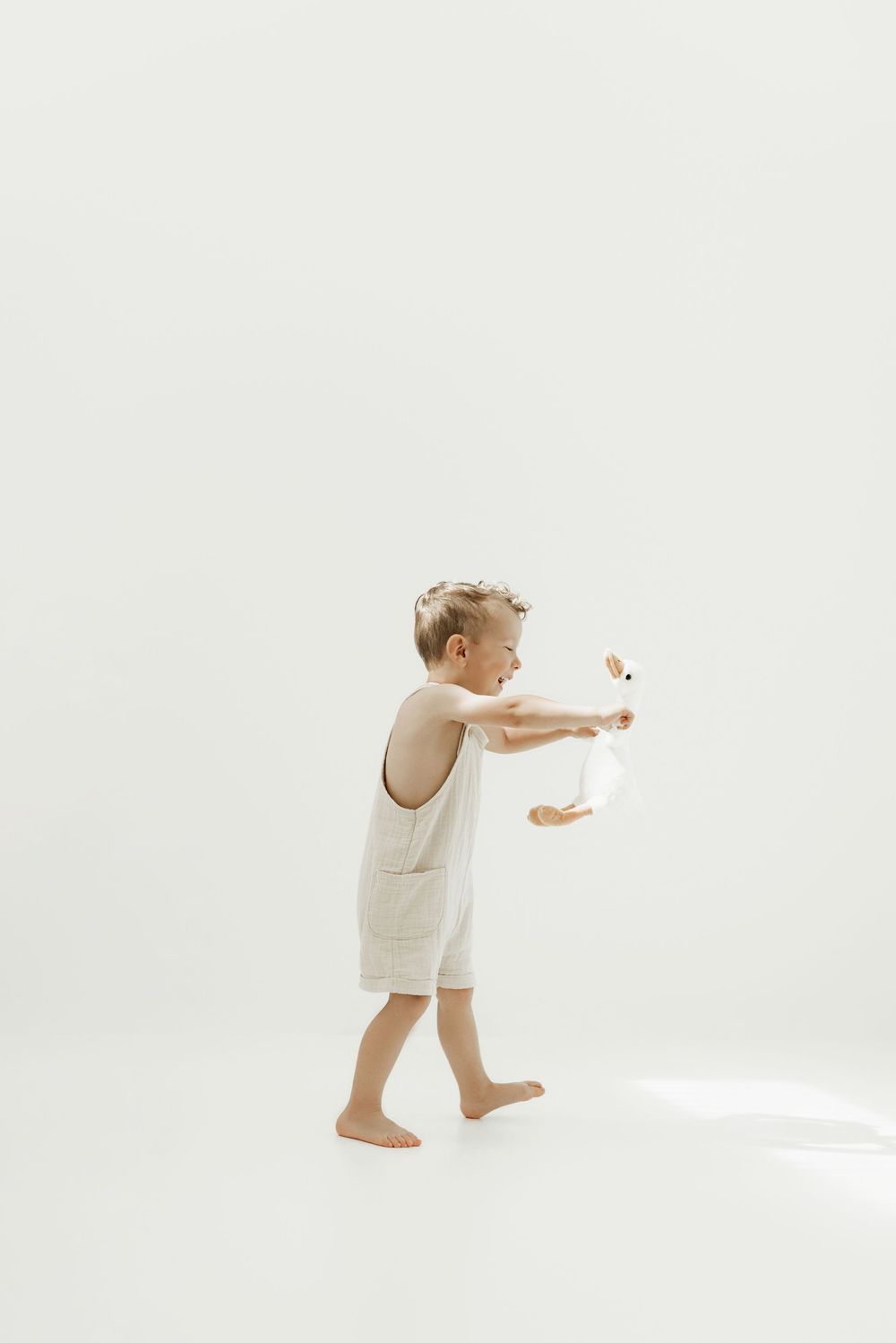Small child in white romper reaching out while walking barefoot against minimal white background.