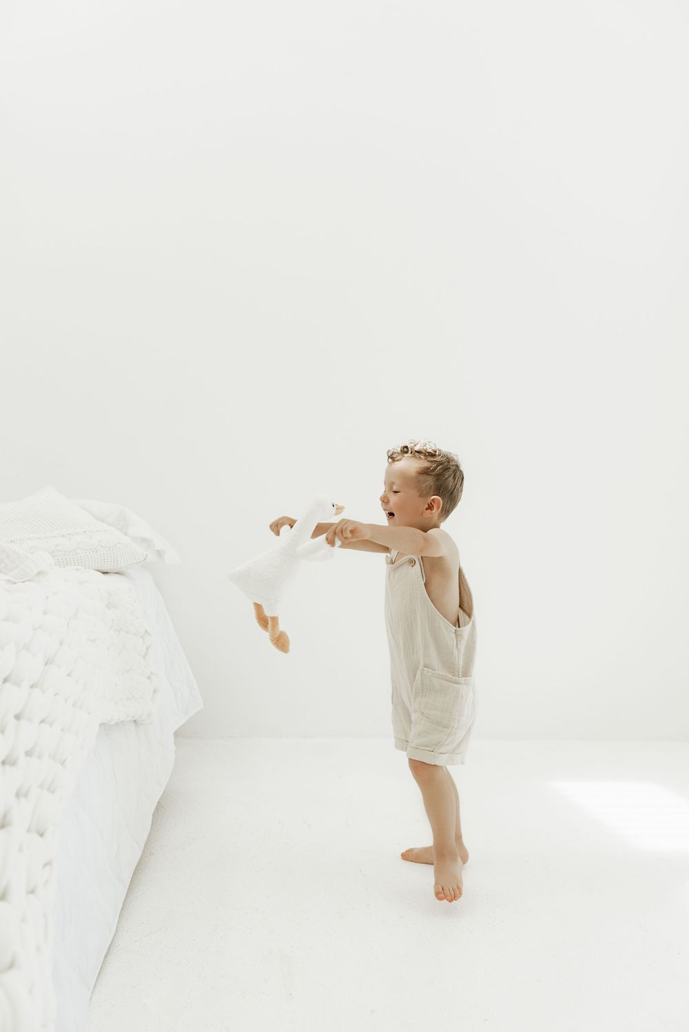 A toddler in white clothing stands on tiptoe reaching towards a white bed in a bright minimalist room.