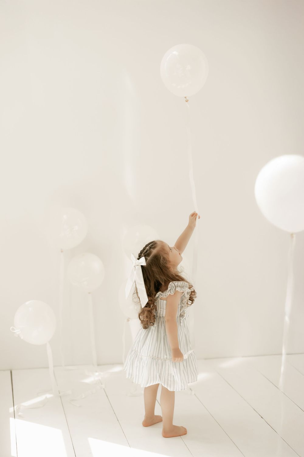 A small figure in a white striped dress reaches up toward white balloons in a bright minimalist setting.