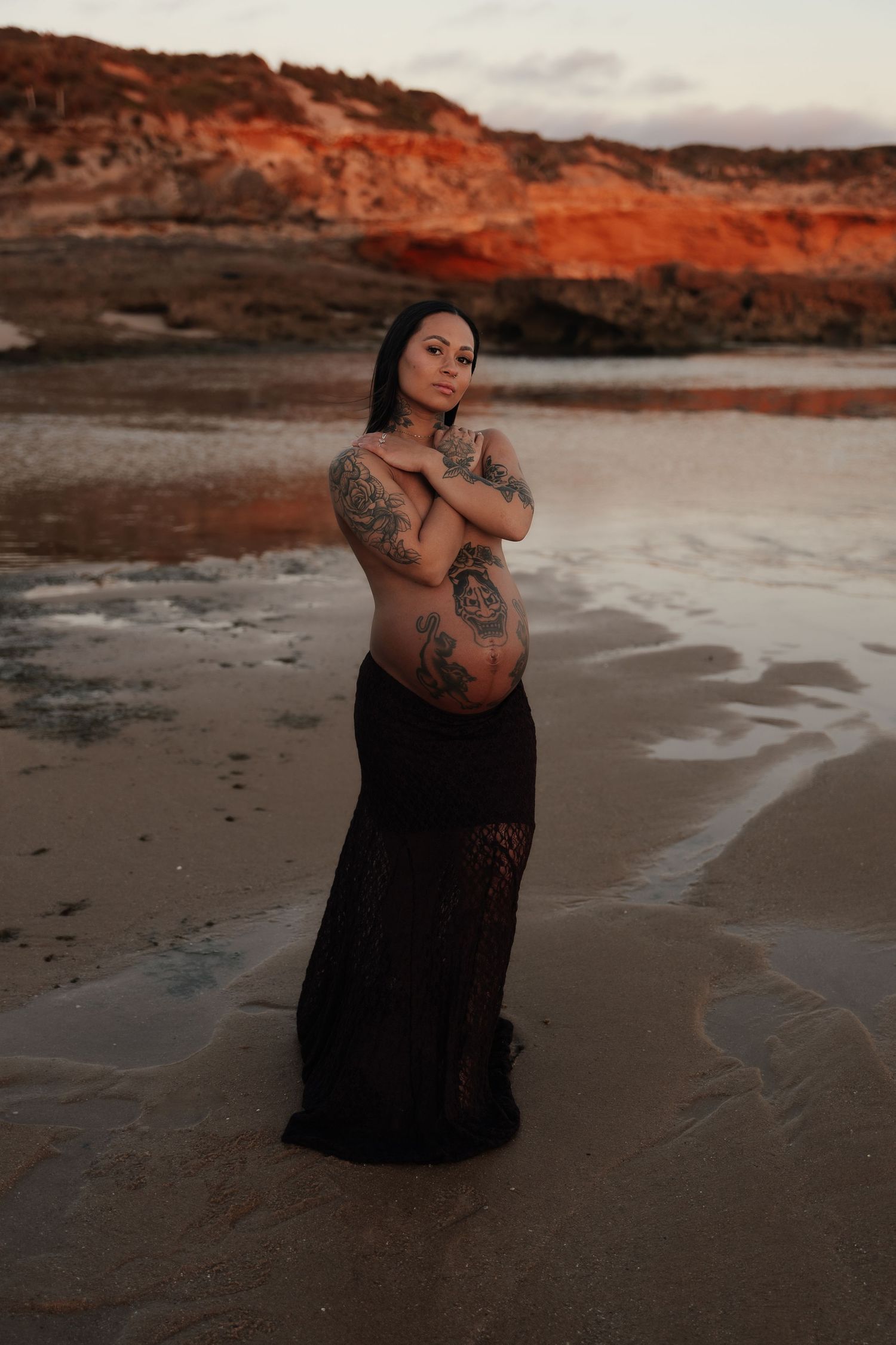 Person in white lingerie and jeans poses on a beach at sunset with dramatic coastal rock formations in the background.
