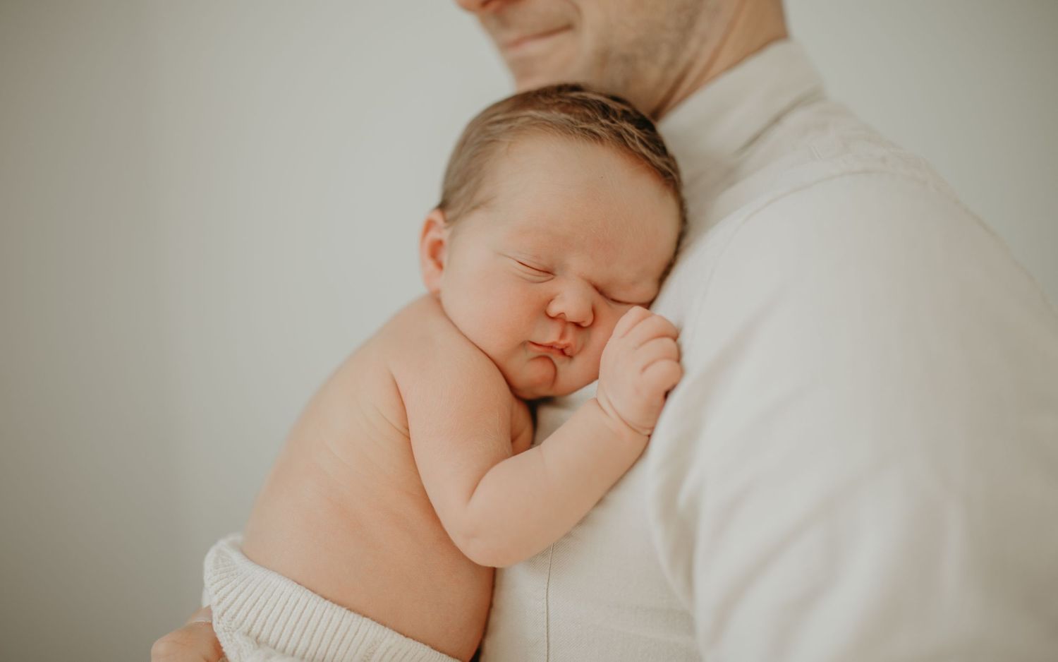 A sleeping newborn baby in a white diaper rests peacefully against a caregiver wearing light colored clothing.