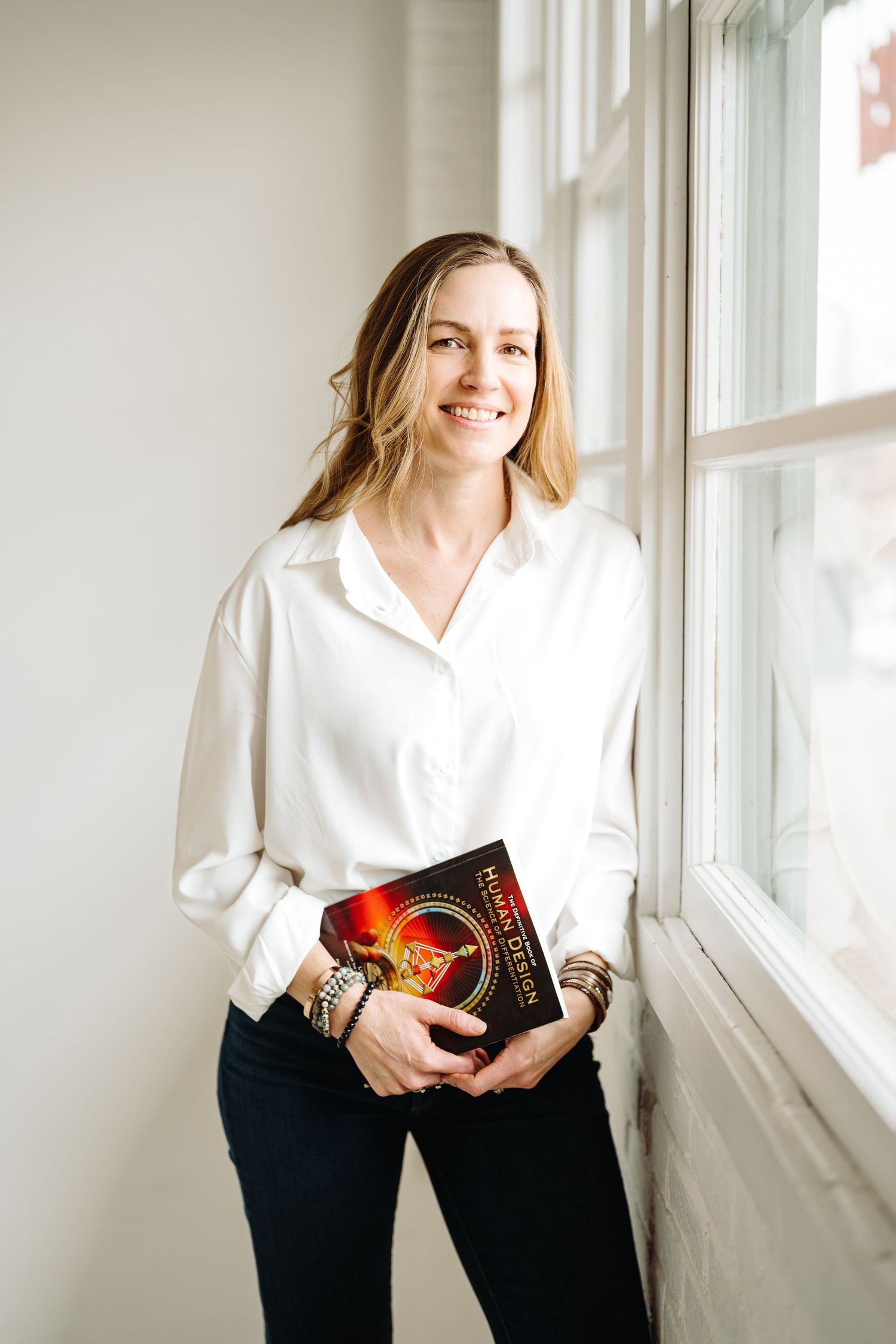 A person in a white shirt and black pants poses with a book near bright windows in a minimalist interior setting.