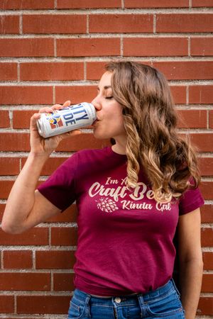 Person in burgundy t-shirt with logo drinking from a can against brick wall background. photography st louis.