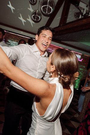 A couple in white outfits share a joyful moment dancing together at a party under decorative lighting  at Birtsmorten Court