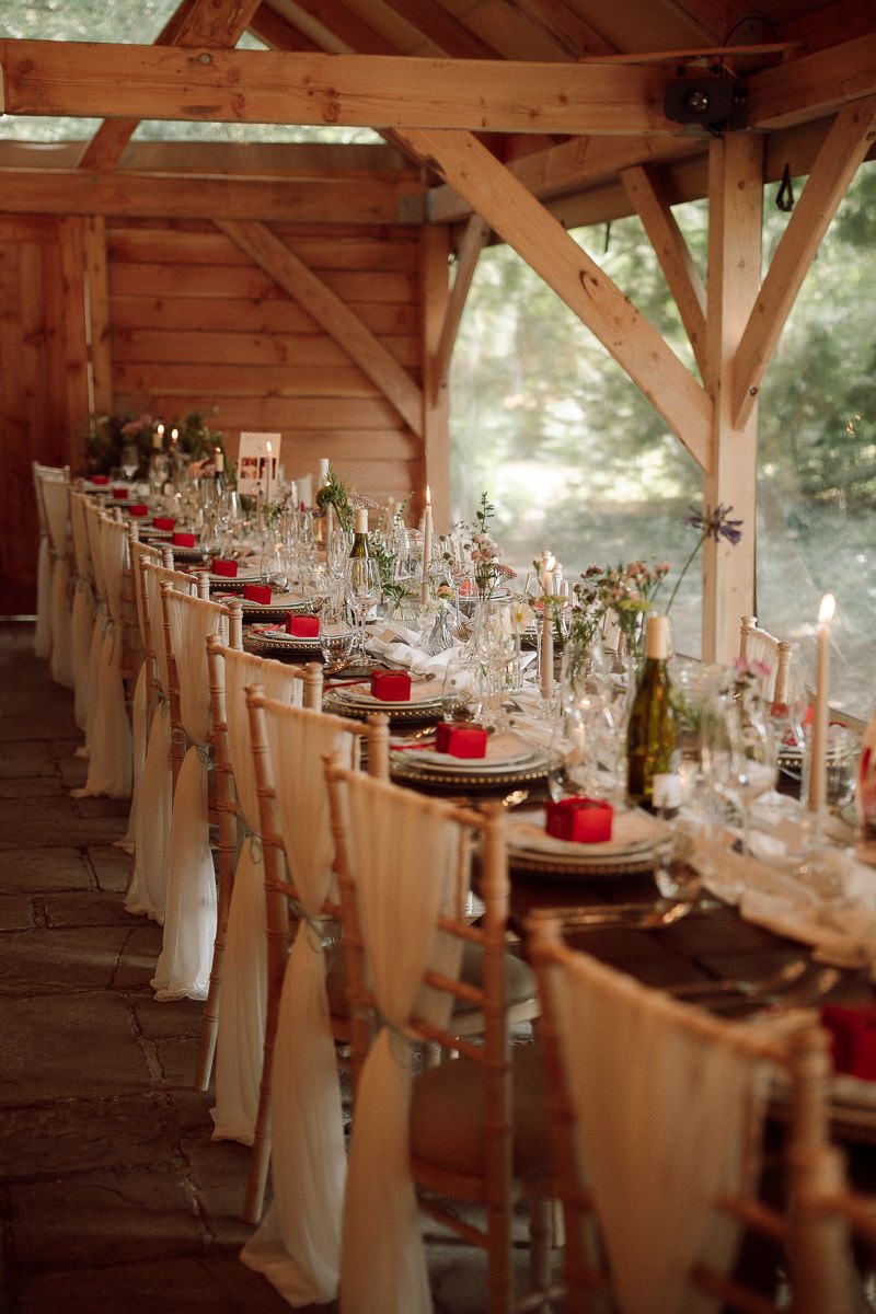 Long rustic wooden tables decorated with elegant glassware, white linens, and red candles under  timber-framed pavilion at Birtsmorten Court
