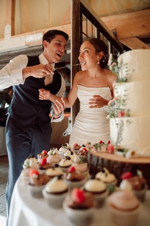 A couple in wedding attire shares a playful moment while cutting cupcakes at their reception  at Birtsmorten Court
